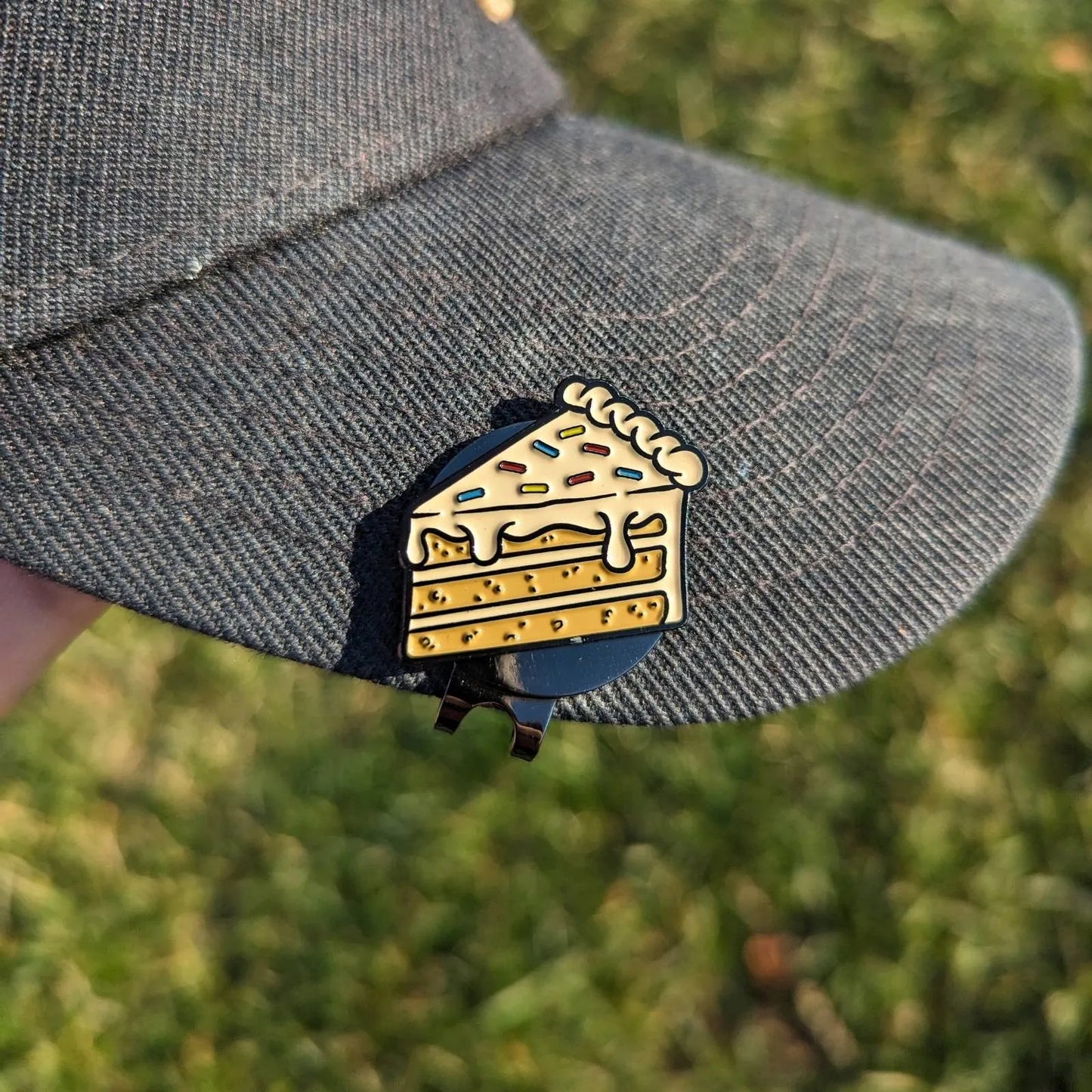 cake golf ball marker with white frosting and rainbow sprinkles clipped on a hat